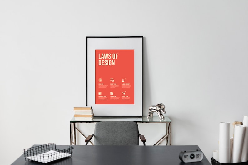 May include: A framed poster with a red background and white text that reads "Laws of Design". The poster is hanging on a white wall above a desk with a black surface. There are books, a silver elephant figurine, and a black alarm clock on the desk.
