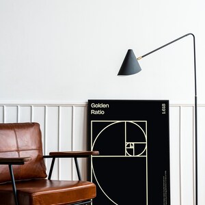 May include: A black and white poster with the text "Golden Ratio" and a geometric design of overlapping circles. The poster is hanging on a white wall next to a brown leather armchair and a black floor lamp.