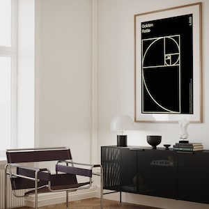 May include: A black and white poster with the text "Golden Ratio" and a geometric design in gold. The poster is framed in a light wood frame and hanging on a white wall. A black cabinet with a grid pattern is in the foreground.