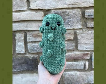 May include: A green crocheted pickle plush toy with a smiling face and black eyes. The toy is being held in a hand against a brick wall background. The text "Pattern By @littlekaycrochet" is at the bottom of the image.