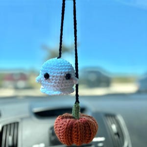 May include: A white crocheted ghost and an orange crocheted pumpkin hanging from a rearview mirror in a car.