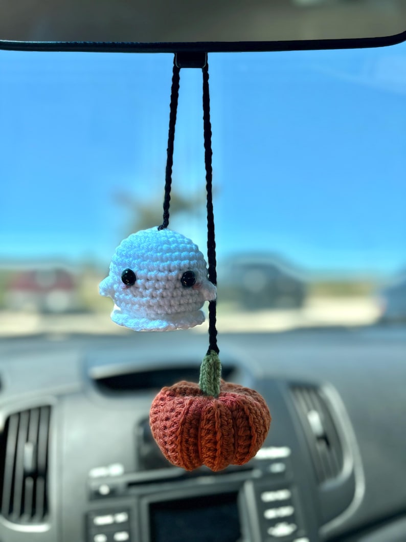 May include: A white crocheted ghost and an orange crocheted pumpkin hanging from a rearview mirror in a car.