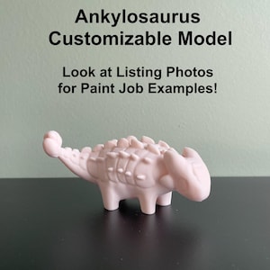 May include: A pink, 3D printed Ankylosaurus dinosaur model. The model is customizable and has a textured, bumpy surface. The text "Ankylosaurus Customizable Model" and "Look at Listing Photos for Paint Job Examples!" is visible in the image.