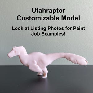 May include: A pink 3D printed Utahraptor dinosaur model. The model is posed in a running position with its tail extended. The text "Utahraptor Customizable Model" and "Look at Listing Photos for Paint Job Examples!" is visible above the model.