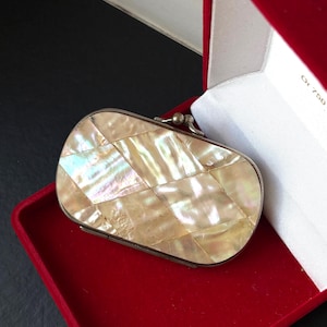 Pode incluir: Um estojo compacto de formato oval com um mosaico de peças de madrepérola iridescente em padrão de losangos. O estojo possui uma moldura metálica prateada e um pequeno fecho esférico, apresentado dentro de uma caixa forrada a veludo vermelho.