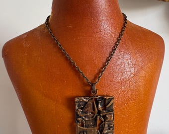 May include: A vintage bronze-toned pendant necklace. The square pendant features an abstract, textured design. The necklace is displayed on a mannequin with a brown leather-like finish. The chain is a matching bronze color.