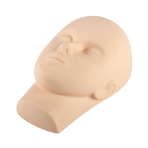 Op de afbeelding: Een beige, levensgrote mannequinhoofd met gesloten ogen, een duidelijke neus en een licht geopende mond. Het hoofd is opzij gekanteld, met een glad oppervlak en een nekbasis. Het oor is zichtbaar aan de zijkant van het hoofd.