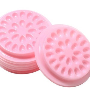 Könnte beinhalten: Ein Satz aus fünf rosa Plastikpaletten mit mehreren kleinen Vertiefungen zum Aufbewahren von Wimpernkleber oder anderen Klebstoffen. Jede Palette hat ein kreisförmiges Design mit einer erhöhten Mitte.