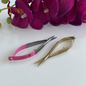 Könnte beinhalten: Zwei Paare kleiner, scharfer Scheren. Ein Paar ist silberfarben mit einem rosa Griff und dem Text "Shiakidi" darauf. Das andere Paar ist golden.