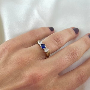 Op de afbeelding: Een gouden ring met vijf stenen. De middelste steen is een grote, donkerblauwe saffier. De andere vier stenen zijn kleiner en helder.