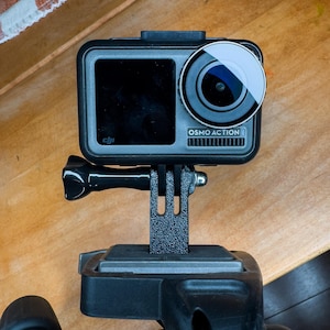 May include: A black and silver DJI Osmo Action camera mounted on a tripod with a black handle. The camera has a small screen on the back and a large lens on the front. The text "OSMO ACTION" is visible on the front of the camera.