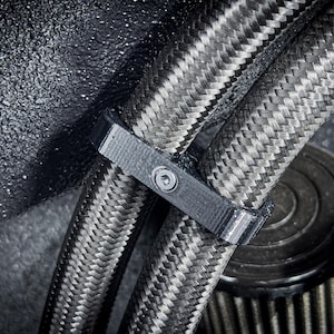 May include: Close-up of two black braided hoses secured with a black plastic clamp. The hoses have a woven texture and are secured together with a screw.