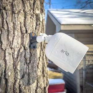 May include: A white Arlo security camera mounted on a tree trunk. The camera is covered in frost and has a black mounting bracket.