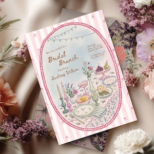 Puede incluir: Una tarjeta de invitación rectangular con un borde de rayas rosas y blancas que presenta una ilustración ovalada de una mesa puesta para un brunch nupcial. El texto dice: You are invited to a Bridal Brunch honoring Audrey Wilson, Sunday June 20, 2027, start 3:00 PM, 123 Anywhere St. Any City 12345.