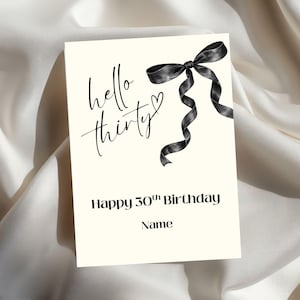 Puede incluir: Tarjeta de cumpleaños color crema con el texto "hello thirty" y "Happy 30th Birthday" en escritura negra. Un lazo negro con cintas que fluyen está en la esquina superior derecha. La tarjeta está sobre una superficie de satén blanco.