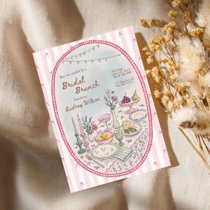 May include: A rectangular invitation card with a pink and white striped border featuring small bows. The center oval illustration depicts a table set for a Bridal Brunch for Audrey Wilson, including candles, flowers, fruit, and plates of food.