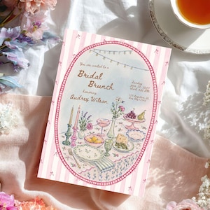 May include: A rectangular invitation card with a pink and white striped border featuring a watercolor illustration of a table set for a bridal brunch. The text reads: You are invited to a Bridal Brunch honoring Audrey Wilson, Sunday June 20, 2027, 3:00 PM, 123 Anywhere St. Any City 12345.