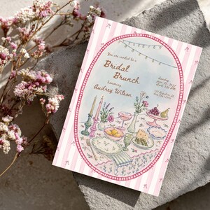 May include: A rectangular invitation card featuring a pink and white striped border with small bow illustrations. The center oval graphic depicts a table setting with candles, flowers, fruit, and plates, with text reading Bridal Brunch honoring Audrey Wilson.