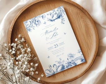 Puede incluir: Una invitación de boda con un diseño de cisnes azules y blancos. La invitación incluye los nombres "Alexandra & Luke" y la fecha "23 de agosto". La invitación está sobre una bandeja de madera con flores blancas.