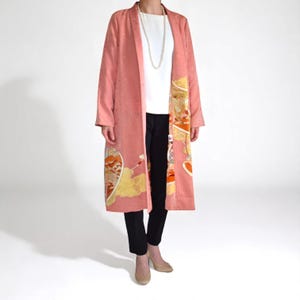 Könnte beinhalten: Ein langer, korallenfarbener Kimono-Stil-Mantel mit dekorativen Paneelen mit orangefarbenen, gelben und goldenen Designs. Der Mantel wird über einem weißen Oberteil und einer schwarzen Hose getragen. Eine lange Perlenkette ist ebenfalls sichtbar.