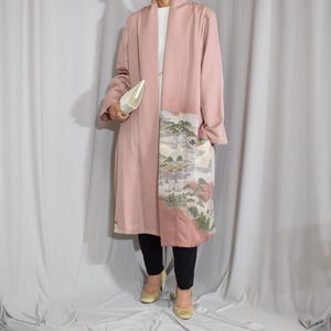 Könnte beinhalten: Ein langer, rosafarbener Kimono-Mantel mit Landschaftsdesign auf der Vorderseite. Der Mantel wird über einem weißen Oberteil und einer schwarzen Hose getragen. Eine weiße Clutch und goldene Absätze vervollständigen den Look. Der Mantel ist ein einzigartiges Kleidungsstück.