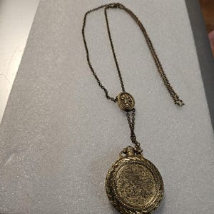Può includere: Una collana vintage color oro con un ciondolo dettagliato. Il ciondolo è di forma ovale con un design intricato e la catena ha un aspetto delicato e antico. La collana presenta un elemento decorativo più piccolo nella parte superiore.