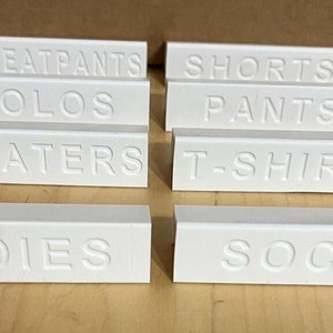 May include: Eight white rectangular labels with different clothing categories written on them. The labels read: "SWEATPANTS", "SHORTS", "POLOS", "PANTS", "SWEATERS", "T-SHIRTS", "UNDIES", and "SOCKS".