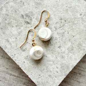 Può includere: Un paio di orecchini pendenti con perline rotonde di conchiglia shiva bianca con motivi a spirale naturali. Ogni perlina è sospesa a un gancio in metallo dorato. Le conchiglie presentano sottili vortici color crema e marrone chiaro sulla superficie.