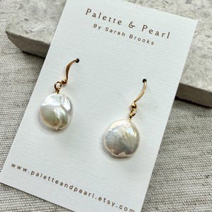 Puede incluir: Un par de pendientes colgantes con perlas blancas de forma irregular suspendidas de ganchos de metal dorado. Los pendientes están sujetos a una tarjeta blanca impresa con el texto Palette & Pearl By Sarah Brooks y la URL del sitio web.