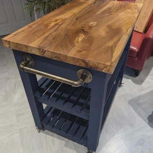 May include: A kitchen island with a dark blue base and a natural wood countertop. The island has a metal towel rail and two open shelves. It is on wheels. The countertop is approximately 100 cm in length.