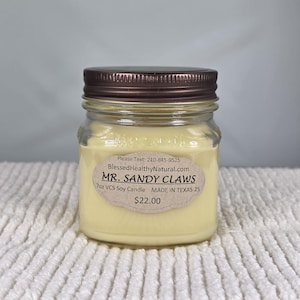 May include: A small, clear glass jar candle with a brown metal lid. The candle is filled with a pale yellow wax. A label on the front reads "MR. SANDY CLAWS" and "7oz VCS Soy Candle MADE IN TEXAS-25".