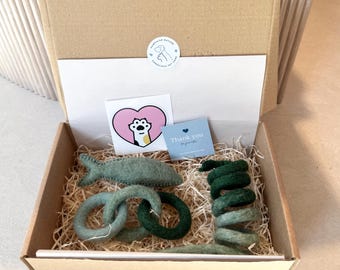 May include: A gift box filled with green felt cat toys, including a fish, a spiral, and rings. The box also includes a thank you card and a sticker with a paw print.