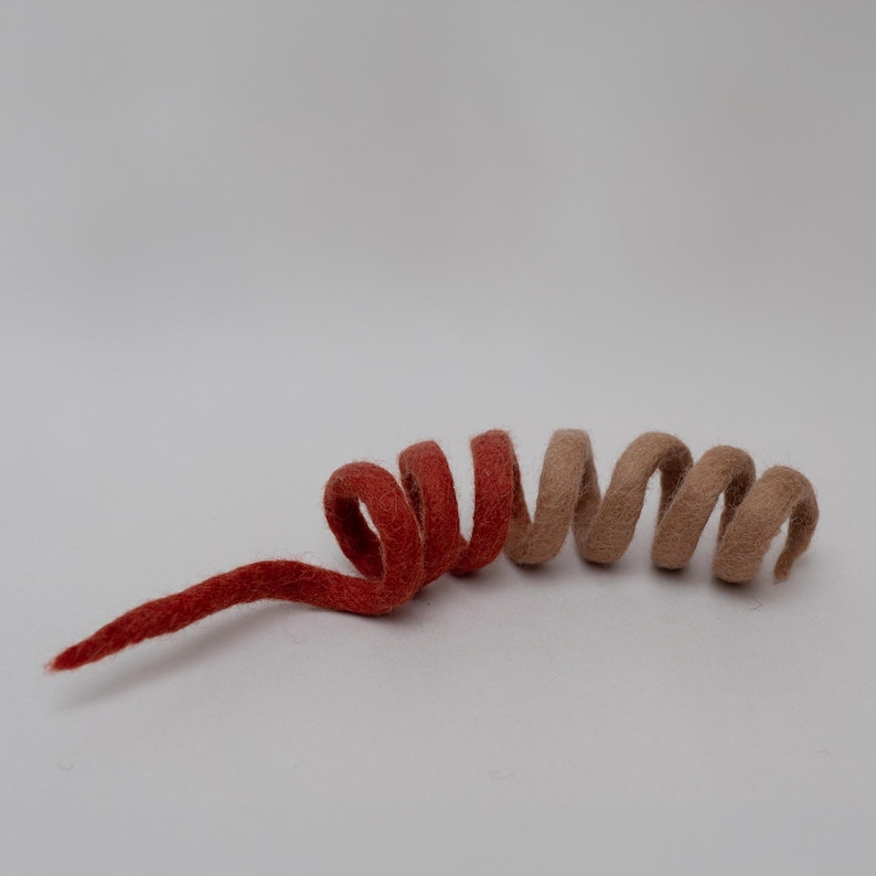 Op de afbeelding: Een spiraalvormige decoratie van gevilt wol met een kleurverloop van diep, verzadigd rood aan de linkerkant naar een zachte, gedempte beige kleur aan de rechterkant. Het object is gevormd in een doorlopende, spiraalachtige krul.