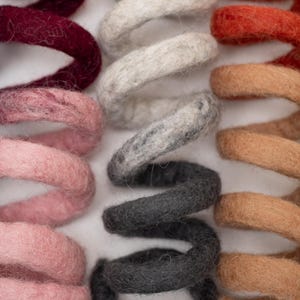 Op de afbeelding: Een verzameling spiraalvormige vilten wollen spiralen gerangschikt in parallelle rijen. De spiralen hebben een verscheidenheid aan kleuren, waaronder blauw, taling, bordeaux, roze, wit, grijs, antraciet, oranje, beige en lichtgroen met een textuur.