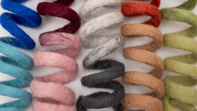Op de afbeelding: Een verzameling spiraalvormige vilten wollen spiralen gerangschikt in parallelle rijen. De spiralen hebben een verscheidenheid aan kleuren, waaronder blauw, taling, bordeaux, roze, wit, grijs, antraciet, oranje, beige en lichtgroen met een textuur.