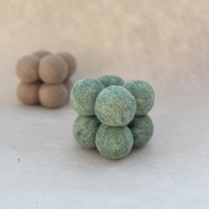 Op de afbeelding: Twee vilten balsculpturen, één in gedempt groen en de andere in een lichtbruine tint. Elke sculptuur is samengesteld uit zeven bollen gerangschikt in een kubusachtige formatie. De vilten ballen hebben een zacht, getextureerd uiterlijk.