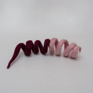 Op de afbeelding: Een spiraalvormige decoratie van gevilt wol met een tweekleurig ontwerp. De linkerkant is diep bordeauxrood en gaat over in een zacht pastelroze aan de rechterkant. Het object is gevormd tot een continue, flexibele spiraal met een pluizige textuur.
