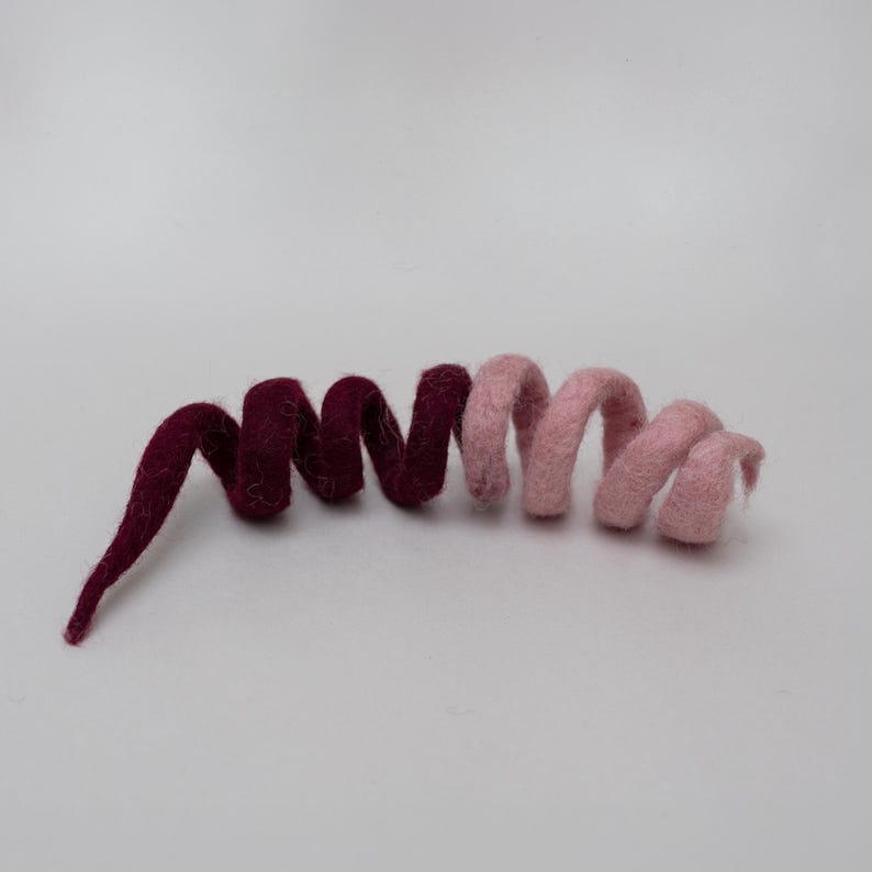 Op de afbeelding: Een spiraalvormige decoratie van gevilt wol met een tweekleurig ontwerp. De linkerkant is diep bordeauxrood en gaat over in een zacht pastelroze aan de rechterkant. Het object is gevormd tot een continue, flexibele spiraal met een pluizige textuur.
