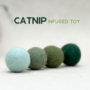 Może przedstawiać: Cztery kulki z filcu nasączone kocimiętką w odcieniach niebieskiego, zielonego i ciemnozielonego. Tekst "CATNIP INFUSED TOY" jest wyświetlany nad kulkami w kolorze ciemnozielonym.