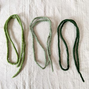 Könnte beinhalten: Drei grüne Filzwollschnüre in verschiedenen Grüntönen. Die Schnüre sind geschlungen und vor einem weißen, strukturierten Hintergrund angeordnet. Die Farben reichen von hell- bis dunkelgrün.