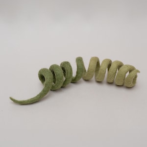 Puede incluir: Una espiral de fieltro verde hecha a mano. El fieltro presenta una mezcla de tonos verdes claros y oscuros, creando un efecto degradado. La forma en espiral recuerda a un zarcillo de planta o un elemento decorativo.