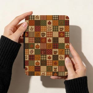 May include: A rectangular e-reader cover featuring a patchwork design of autumn-themed squares. The pattern includes orange pumpkins, brown coffee mugs, red and gold maple leaves, and various plaid, polka dot, and knit-textured fabric prints.