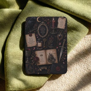 May include: A rectangular e-reader cover featuring a dark, gothic-inspired collage of a raven, a crescent moon, a burning candle, a dagger, a goblet, a key, a potion bottle, and an open book with text, all set against a black background.