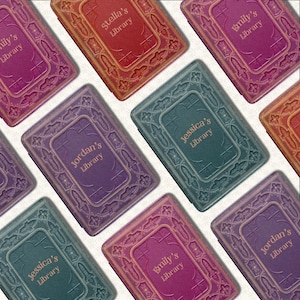 May include: An assortment of personalized library boxes in various colors, including red, purple, and teal. Each box is embossed with a decorative border and features a name and the word "Library" in a serif font.