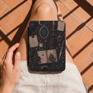 May include: A black e-reader case featuring a dark, gothic-inspired pattern with illustrations of a candle, a dagger, an open book with a castle, a potion bottle, a key, and various text snippets including "I would burn the world for you."