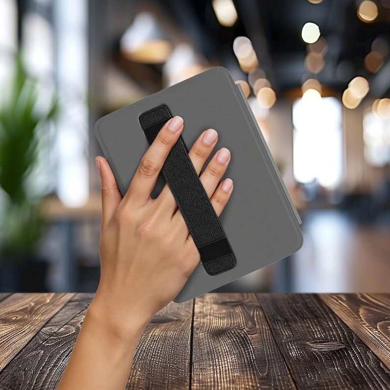 May include: A charcoal gray tablet case featuring a black elastic hand strap attached diagonally across the back. The case has a smooth, matte finish and a rectangular shape with rounded corners, designed to provide a secure grip for the device.