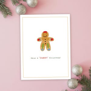 May include: A white card with a gingerbread man illustration and the text "Have a 'SWEET' Christmas!"  The card is surrounded by silver ornaments and pine branches on a pink background.