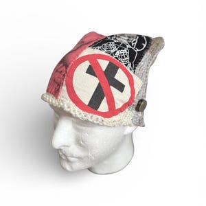 May include: A multi-panel beanie constructed from various fabric patches, featuring a prominent red circle with a diagonal line over a black cross on a cream background, a black and white graphic of a skeleton, and a pink section with a metallic button.