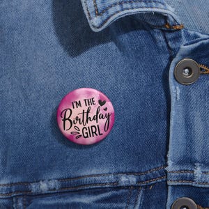 May include: A pink and white button with the text "I'm the Birthday Girl" in black lettering and two small black hearts.