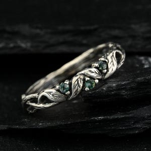 May include: A silver ring featuring a band designed with sculpted leaves and vines. The metal has an oxidized finish to highlight the intricate textures. Three small, round, green moss agate gemstones are set within the leaf-patterned band.